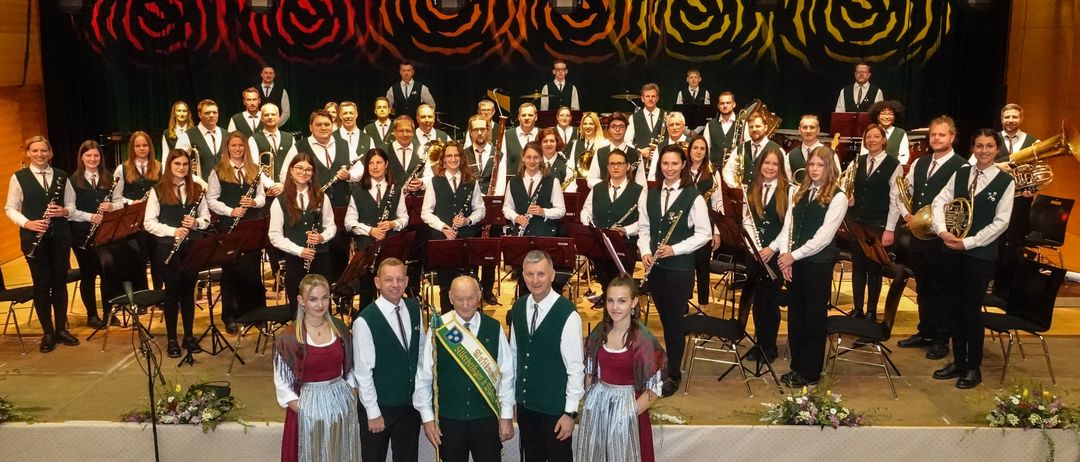 Ein großes Ensemble von Musikern steht auf der Bühne, einige halten Instrumente. Vorne stehen mehrere Personen, und zwei Frauen tragen traditionelle Kleider. Die Bühne ist mit Blumen und Scheinwerfern geschmückt.