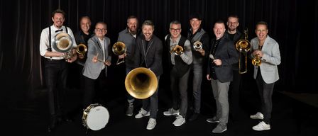 Eine Gruppe von Musikern, alle mit Blechblasinstrumenten, posiert für ein Foto mit einem schwarzen Vorhang als Hintergrund.