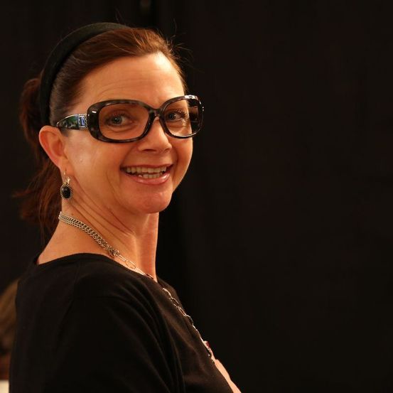 Eine Frau, die für ein Foto lächelt, trägt eine Brille, eine schwarze Kopfbinde, eine Halskette und Ohrringe. Sie hat kurze Haare und trägt ein schwarzes Oberteil.