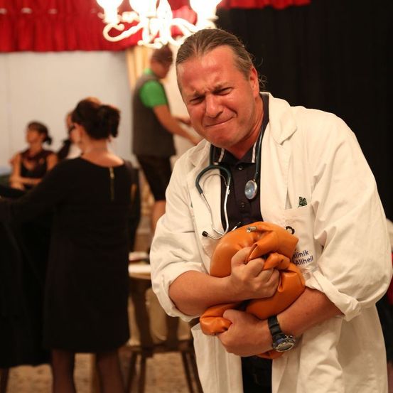 Ein Arzt hält ein orangefarbenes Objekt in den Händen, möglicherweise ein medizinischer Dummy, mit einem Stethoskop um den Hals.