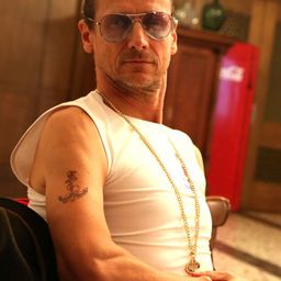 Ein Mann mit Sonnenbrille und einem ärmellosen weißen T-Shirt sitzt in einem Stuhl und trägt eine goldene Halskette mit Anhänger. Er hat eine Tätowierung eines Ankers auf seinem Arm. Hinter ihm steht ein Holzschrank mit einem Coca-Cola-Kühler.