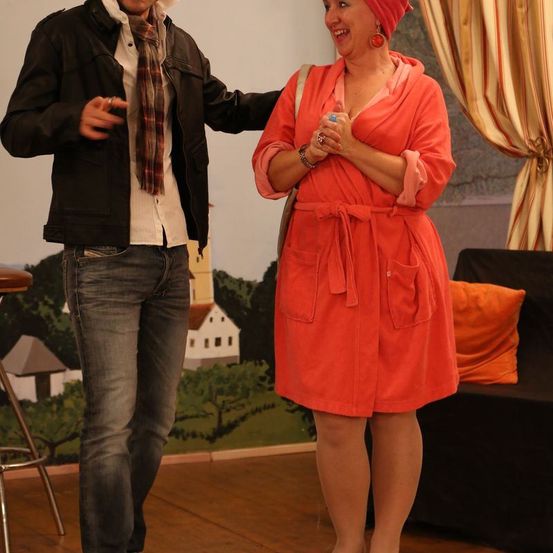 Eine Frau in einem leuchtend orangefarbenen Bademantel und ein Mann in einer schwarzen Jacke und Jeans stehen lächelnd drinnen.