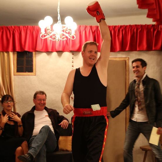 Ein Mann in einem schwarzen Tanktop und roten Boxhandschuhen steht triumphierend mit einem erhobenen Arm und lächelt. Hinter ihm sitzen zwei Personen auf einer Bank und schauen zu, eine mit einem Papier und die andere mit Brille. Eine Lampe hängt über ihnen.