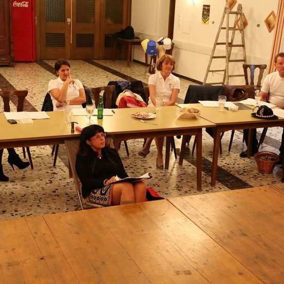 In einem Raum mit langen Tischen, Stühlen und sitzenden und stehenden Personen findet ein Treffen statt. Eine Person schreibt, und andere beobachten.