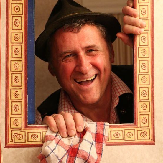 Ein Mann mit schwarzem Hut und rot-weiß kariertem Hemd lächelt, während er einen Spiegelrahmen mit Blumenmustern hält.