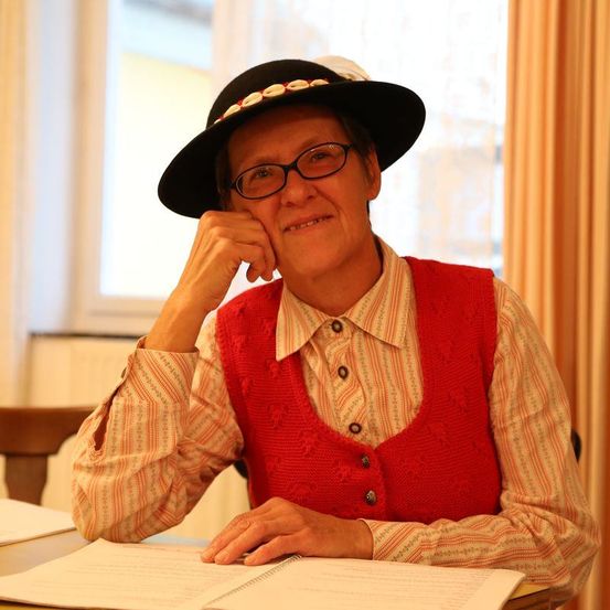 Eine ältere Frau mit Brille und Hut sitzt an einem Schreibtisch mit einem Notizbuch, lächelt und stützt ihren Kinn auf die Hand.