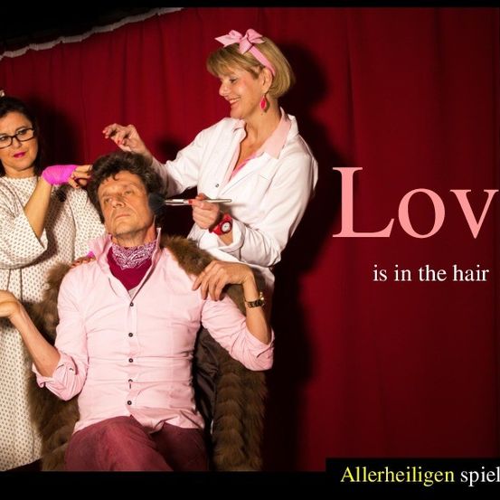 Ein Mann sitzt in einem Stuhl und lässt sich von zwei Frauen die Haare stylen. Eine hält einen Kamm und die andere einen Haartrockner. Der Schauplatz ist eine Bühne mit einem roten Vorhang. Die Worte 'Love is in the hair' erscheinen auf dem Bild.