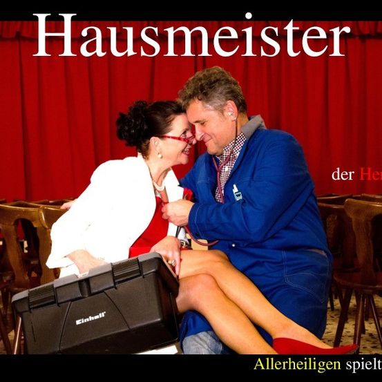 Ein Mann und eine Frau, möglicherweise Schauspieler, sitzen auf einer Bühne. Die Frau hält eine Werkzeugkiste, und der Mann hat ein Stethoskop. Der Hintergrund ist ein roter Vorhang.