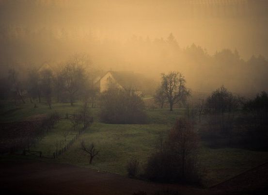 Eine neblige Landschaft mit einem Haus und Bäumen in der Ferne unter einem gelben Himmel.