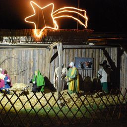 Eine Krippe ist bei Nacht im Freien aufgebaut, mit einem sternförmigen Licht darüber. Mehrere Personen sind in Kostümen gekleidet und stehen in einem Holzzaun.
