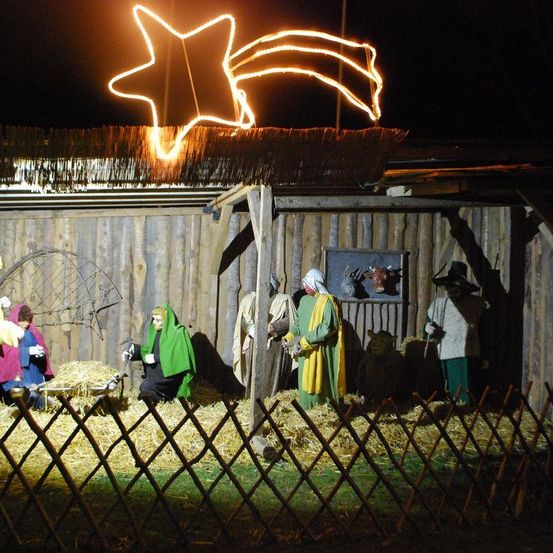 Eine Krippe ist bei Nacht im Freien aufgebaut, mit einem sternförmigen Licht darüber. Mehrere Personen sind in Kostümen gekleidet und stehen in einem Holzzaun.