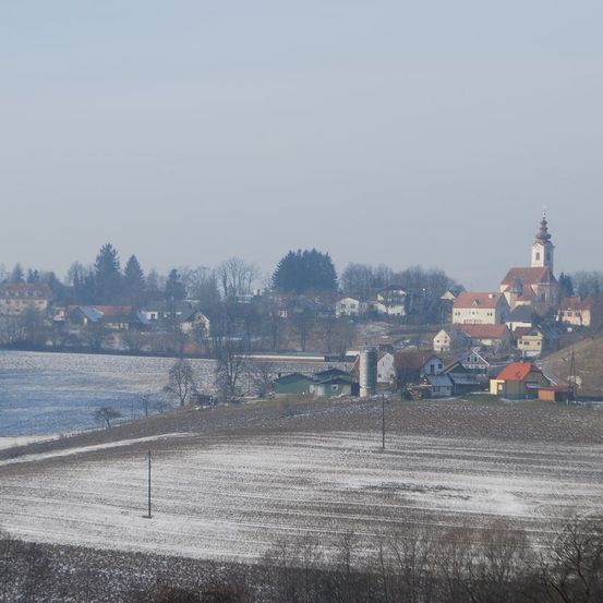 Eine Winterlandschaft zeigt eine Kirche mit einem Turm auf einem Hügel, umgeben von schneebedeckten Feldern und Häusern, mit einem Fluss und Bäumen in der Ferne.