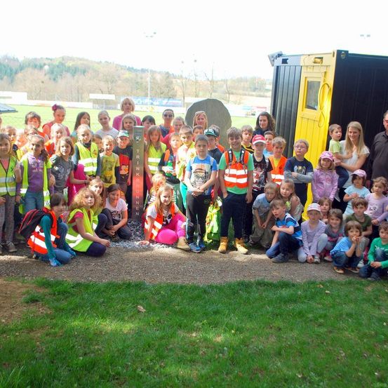 Eine Gruppe von Kindern und Erwachsenen steht in einem Park und trägt Warnwesten. Hinter ihnen ist ein gelber und schwarzer Container zu sehen, und in der Ferne sind ein Bergrücken und Straßenlaternen zu erkennen.