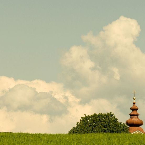 Eine ländliche Szene zeigt eine Kirche mit einer Kupferkuppel unter einem teilweise bewölkten Himmel, umgeben von üppigen grünen Feldern und einem Baum im Vordergrund.