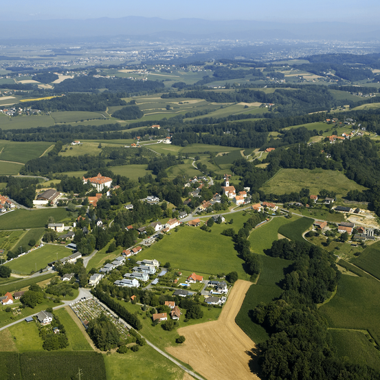 Luftaufnahme eines Dorfes mit zahlreichen Gebäuden, grünen Feldern und Bäumen in einer ländlichen Landschaft mit entfernten Bergen.