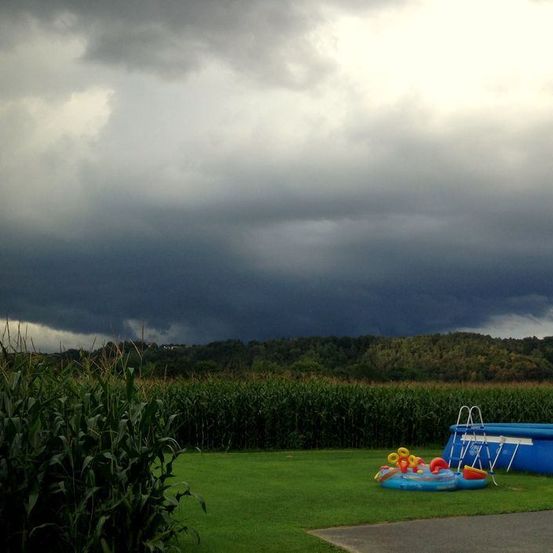 Ein dunkler Gewitterwolke hängt über einem Rasen mit einem aufblasbaren Pool und Spielzeug, in der Nähe eines Maisfeldes mit Bäumen im Hintergrund.