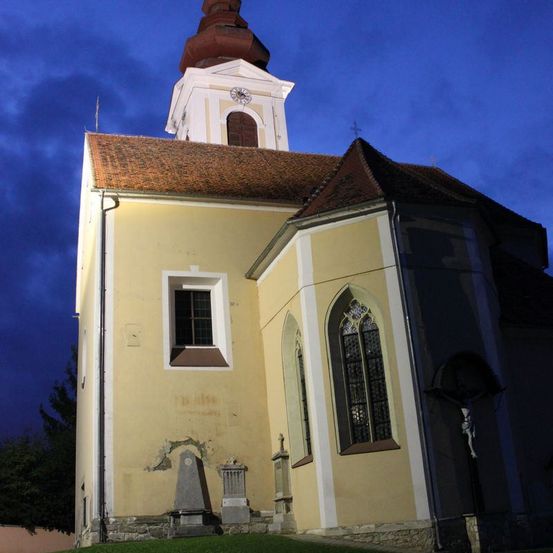 Eine Kirche mit Turm und Dachreiter ist gegen einen dunkelblauen Himmel beleuchtet. Das Gebäude hat ein braunes Dach und eine Uhr auf dem Turm.