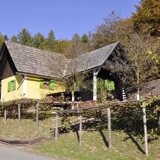 Ein kleines gelbes Haus mit grünen Fensterläden steht neben einer Straße, umgeben von einem Holzzaun und Weinranken. Bäume und Sträucher im Hintergrund.