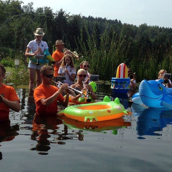 Eine Gruppe von Menschen steht in einem See und spielt Musikinstrumente wie Trompeten und Klarinetten. Einige sitzen auf aufblasbaren Schwimmern. Die Umgebung ist gefüllt mit Bäumen und Pflanzen.