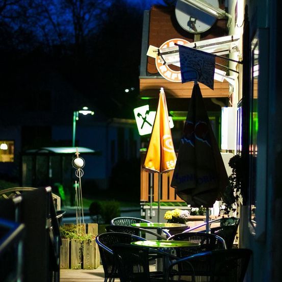 Es wird ein Außenbereich eines Restaurants bei Nacht gezeigt. Die Tische sind von Stühlen umgeben, und zwei zusammengeklappte Regenschirme sind an Stangen befestigt.