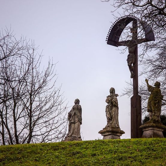 Drei Statuen umgeben ein großes Kruzifix auf einem grasbewachsenen Hügel, mit kahlen Bäumen im Hintergrund.