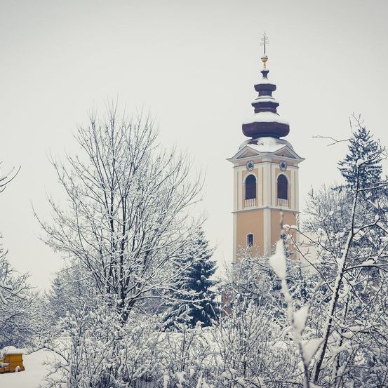 Ein Kirchturm steht inmitten einer verschneiten Winterlandschaft, mit einem gelben Bienenstock im Vordergrund.