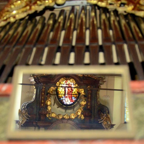 Ein Nahaufnahme von Orgelpfeifen mit der Reflexion eines Buntglasfensters, das eine zentrale Figur in Rot und Gold auf einem hölzernen Rahmen zeigt.