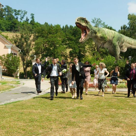 Eine Gruppe von Menschen in Anzügen rennt an einem großen Dinosaurier in einem Park vorbei. Dahinter ist ein Gebäude mit einem großen Baum zu sehen.