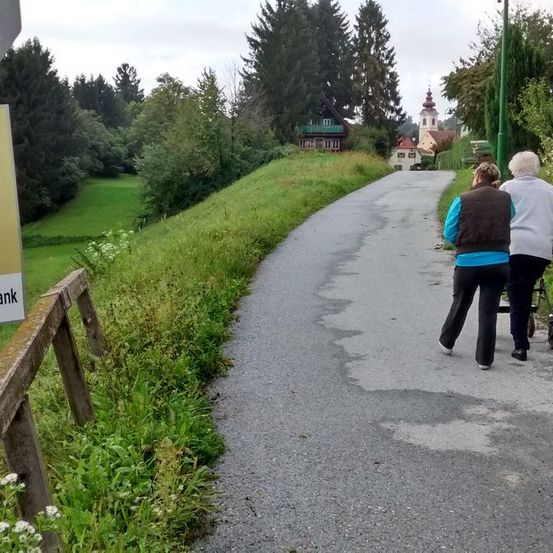 Zwei ältere Frauen gehen einen Schotterweg hinunter, an dem ein Schild mit der Aufschrift 'er' neben einem Holzzaun steht.