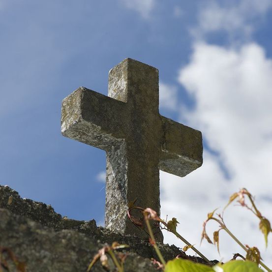 Ein verwittertes Steinkreuz steht auf einem Felsen, umgeben von grünen Pflanzen, vor einem Hintergrund eines klaren blauen Himmels mit einigen Wolken.