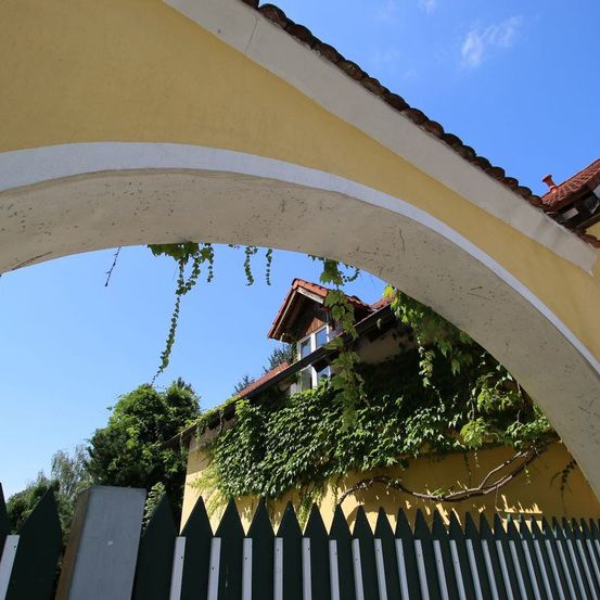 Ein gelbes Haus mit rotem Dach und grünem Efeu, gesehen von unter einem weißen Bogen mit blauem Himmel im Hintergrund.
