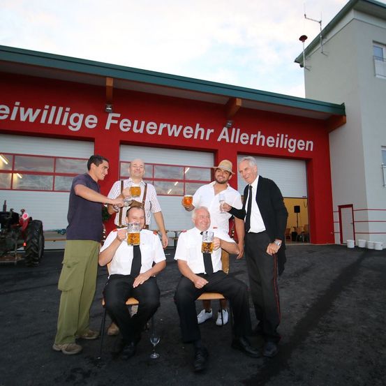 Fünf Männer vor einer Feuerwehrwache mit einem Schild, das Freiwillige Feuerwehr Allerheiligen liest. Drei sitzen, während zwei stehen und alle Biergläser halten.