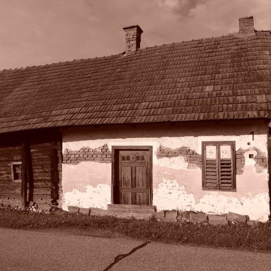 Ein altes, verwittertes Haus mit einer Holztür und zwei Fenstern. Das Dach ist mit Ziegeln gedeckt, und die Wände zeigen Abnutzungsspuren. Das Haus befindet sich an einer Kiesstraße.