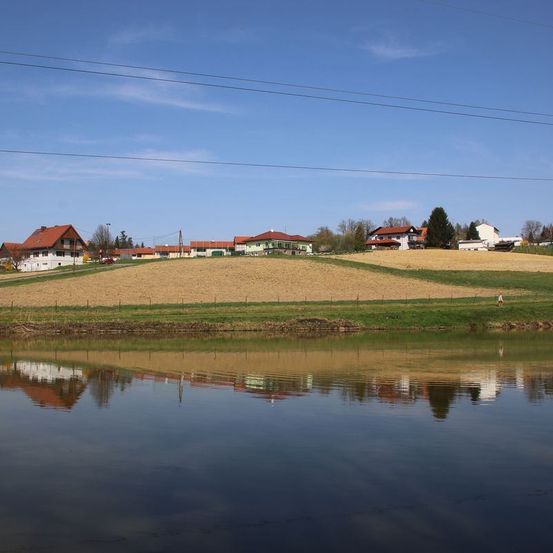 Eine ländliche Landschaft mit Häusern, einem Fluss und Feldern unter einem blauen Himmel. Spiegelung im Wasser.