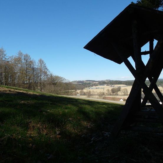 Eine Holzstruktur steht auf einem Grashügel. Sie bietet einen Blick auf ein Tal und Bäume. Der Himmel ist klar und blau.
