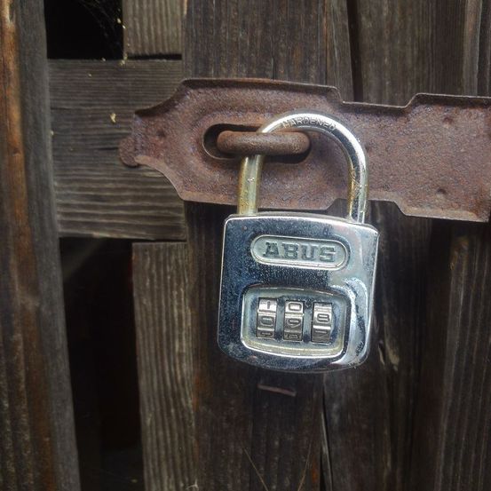 Ein rostiger Vorhangschloss mit Zahlenkombination ist an einer Holzplatte befestigt. Das Schloss trägt die Aufschrift ABUS.