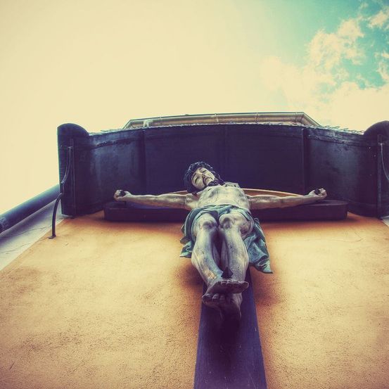 Ein Kruzifix mit Jesus am Kreuz ist an einer Wand montiert. Die Figur ist aus Stein und ruht auf einem Holzbalken.