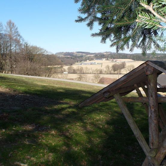 Eine Holzkonstruktion befindet sich in einem Grasfeld. Die Struktur hat ein geneigtes Dach und eine Holzstütze. Es gibt Bäume in der Umgebung, und das Gebiet ist von einem Zaun umgeben. In der Ferne gibt es eine Straße, ein Feld und eine Bergkette. Der Himmel ist klar und blau.