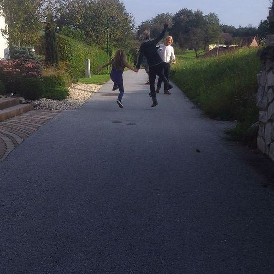 Drei Menschen springen auf einem Weg mit einem Haus und Grünpflanzen im Hintergrund. Das Mädchen springt zuerst, gefolgt von einem Mann und dann einer Frau.