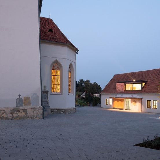 Ein großes Kirchengebäude mit rotem Dach steht in einem Innenhof. Der Eingang ist offen und zeigt ein helles Inneres. Ein weiteres kleineres Haus mit rotem Dach befindet sich in der Nähe.