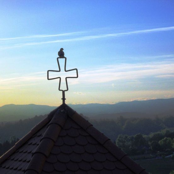 Ein Kreuz auf dem Dach einer Kirche mit einem Vogel, der darauf sitzt. Im Hintergrund ist ein blauer Himmel mit Bergen in der Ferne zu sehen.