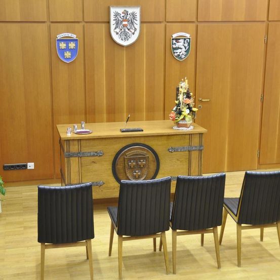 Ein leerer Raum mit Holzmöbeln, einschließlich eines Schreibtisches, Stühlen und einem Wappen an der Wand. Eine Pflanze und ein dekoratives Objekt mit Blumen stehen auf dem Schreibtisch.