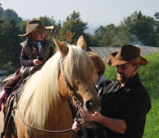 Ein Kind in einem Cowboyhut reitet ein Pferd, während ein Mann an der Leine steht. Eine weitere Frau beobachtet dahinter. Bäume und ein Haus sind im Hintergrund.