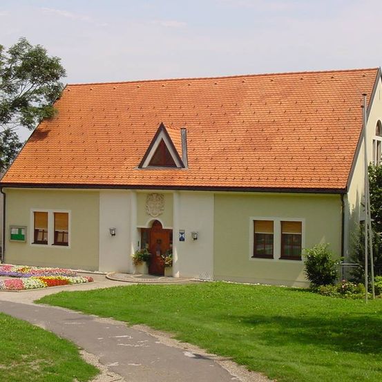 Ein traditionelles Haus mit einem roten Ziegeldach, cremefarbenen Wänden und einer braunen Tür. Es hat ein dreieckiges Fenster über der Tür und zwei kleine Fenster auf beiden Seiten.
