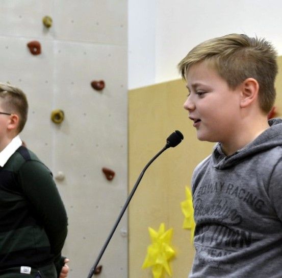 Zwei junge Jungen stehen vor einem Mikrofon, einer scheint zu sprechen, während der andere zuhört, sie befinden sich in einem Raum mit einer Felswand und Sonnendekorationen.