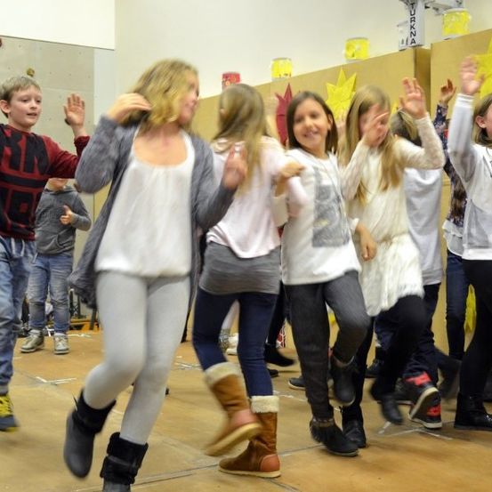 Gruppe von Kindern tanzt auf einer Bühne, einige tragen Stiefel und andere Turnschuhe, mit gelben Sternen an der Wand im Hintergrund.