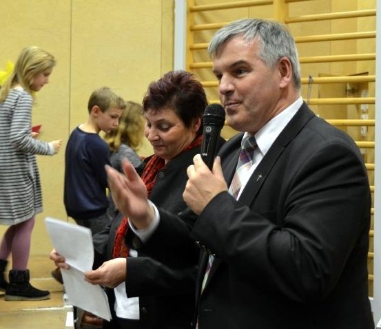 Ein Mann und eine Frau stehen vor einem Mikrofon. Sie klatschen. Kinder stehen hinter ihnen, eines hält ein rotes Objekt. Der Raum hat Holzwände und Ausrüstung.
