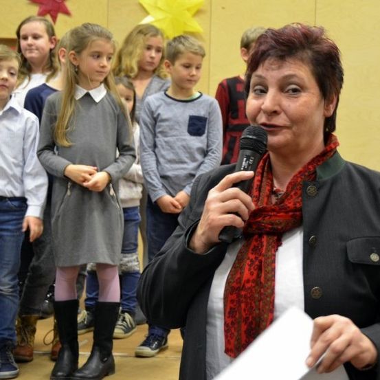 Eine Frau hält ein Mikrofon und steht vor einer Gruppe von Kindern. Sie alle schauen sie an. Sie trägt einen schwarzen Mantel und einen roten Schal. Die Kinder tragen Freizeitkleidung.