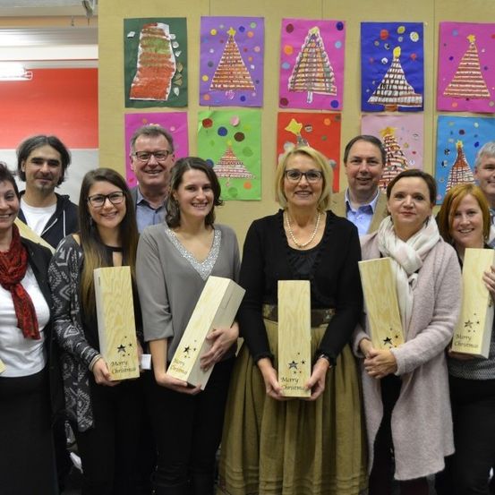 Eine Gruppe von Menschen posiert für ein Foto vor einer Wand mit Weihnachtsbaumgemälden. Sie halten Holzkisten mit der Aufschrift Frohe Weihnachten.