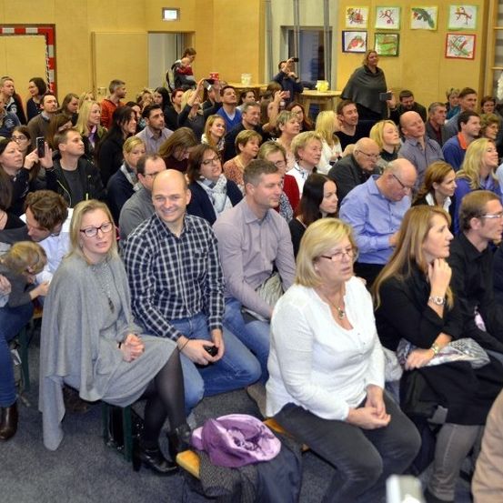 Ein Raum voller Menschen, die einer Veranstaltung beiwohnen. Viele sitzen auf Stühlen, während andere stehen. Einige Personen benutzen ihre Handys, um Fotos oder Videos aufzunehmen. Eine Frau mit einem Kind sitzt mit anderen zusammen. Der Raum hat eine warme Farbgebung und dekorierte Wände.